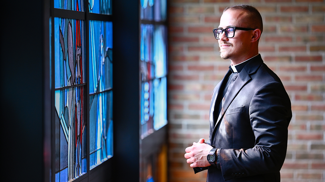 Profile image of a young priest looking out a set of stained glass windows with his hands together
